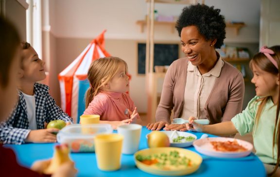 Group of happy kids communicating with their African American teacher while eating lunch at preschool.